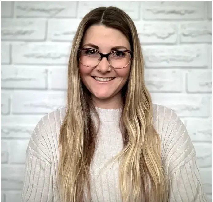 Woman with glasses smiling against a white brick wall.