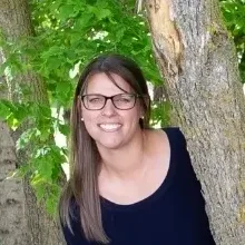 A woman smiling, standing by a tree.