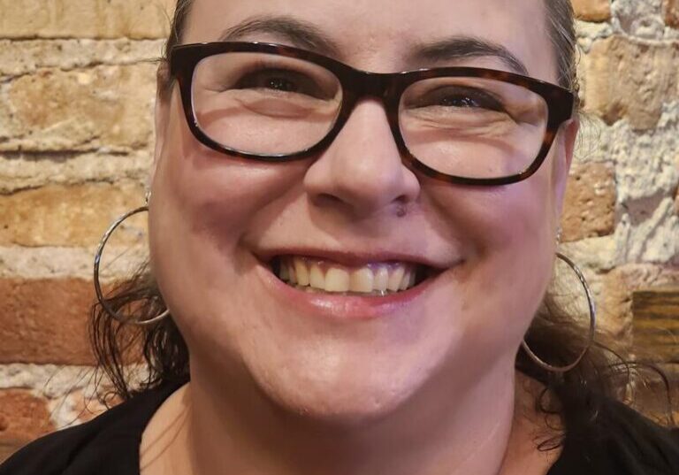Smiling person wearing glasses, brick wall background.