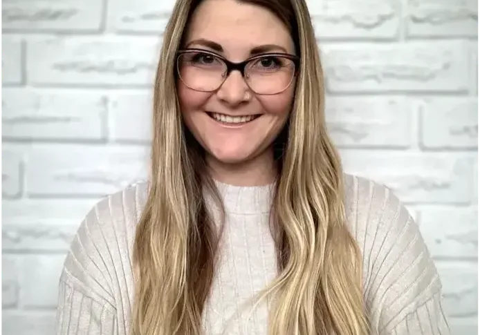 Woman with glasses smiling against a white brick wall.